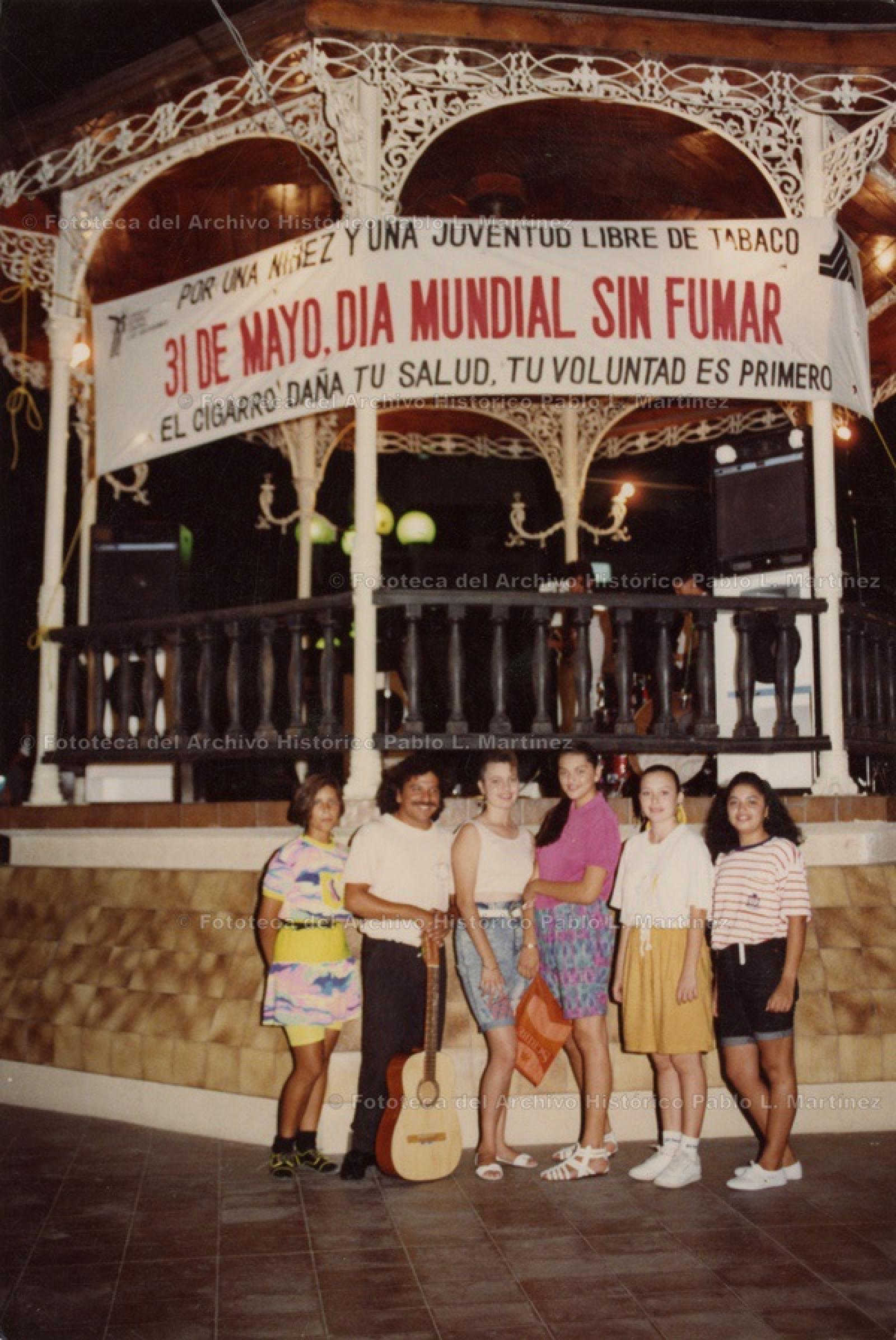 Hombre con guitarra acompañado por grupo de mujeres, frente a kiosco.